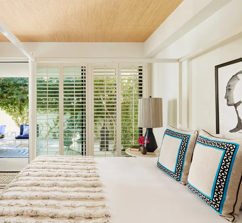 Modern bedroom with a white bed, decorative pillows, fur throw, large window with white shutters, a black and white artwork, lamp, and view of an outdoor patio with greenery.